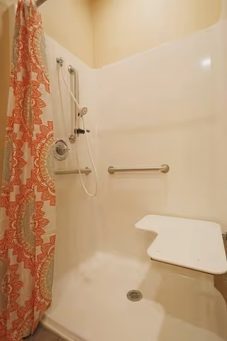 Shower area with a white fold-down seat, grab bars, a handheld showerhead, and a shower curtain with a red and beige pattern.