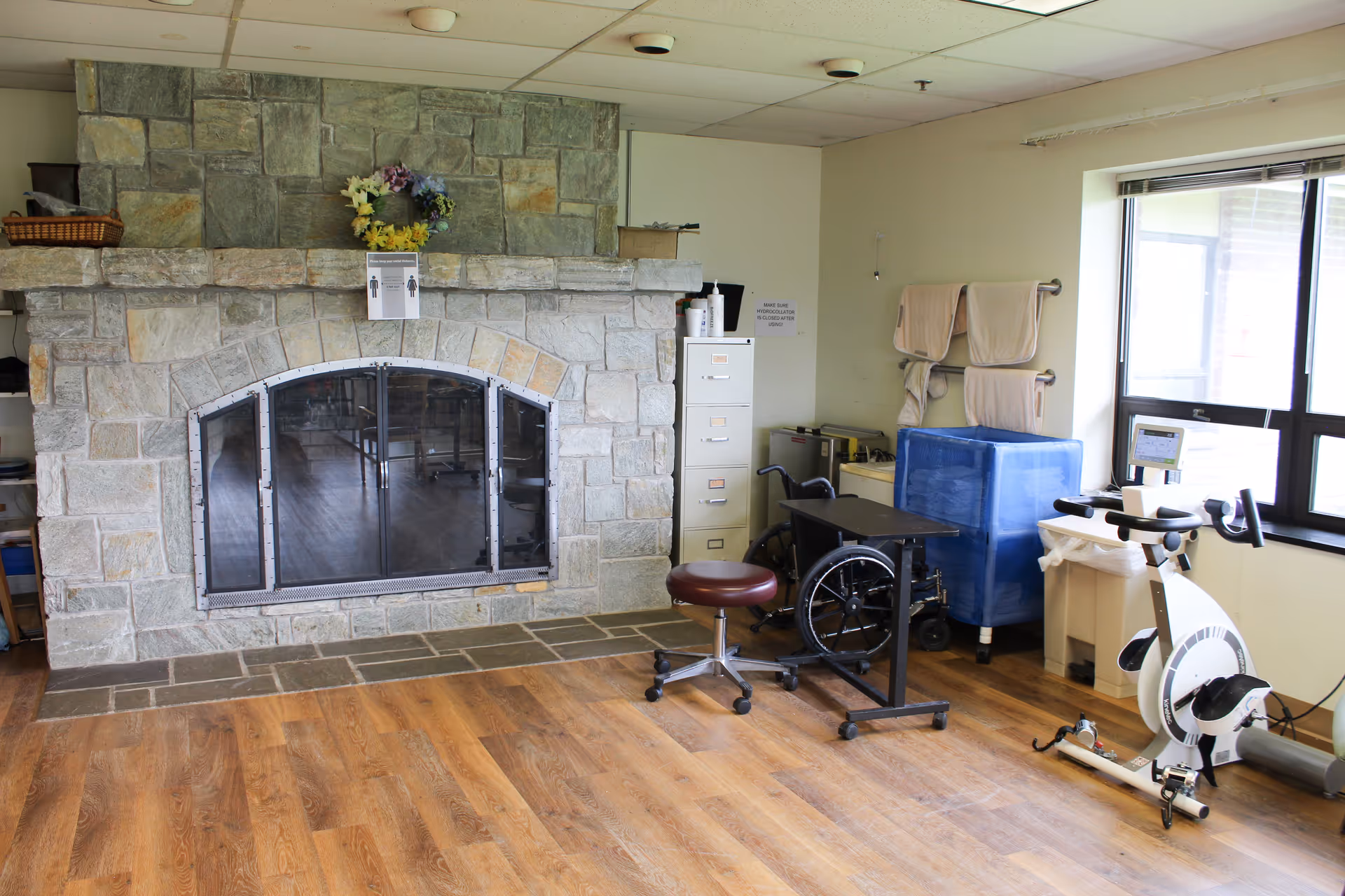 Interior room with a large stone fireplace, wood flooring, an exercise bike and medical equipment near a window.