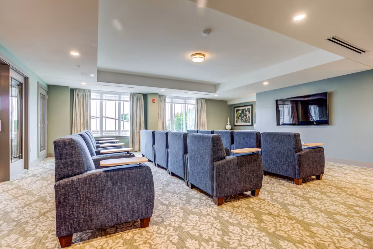Bright common media room with rows of upholstered armchairs facing a wall-mounted TV and large windows.