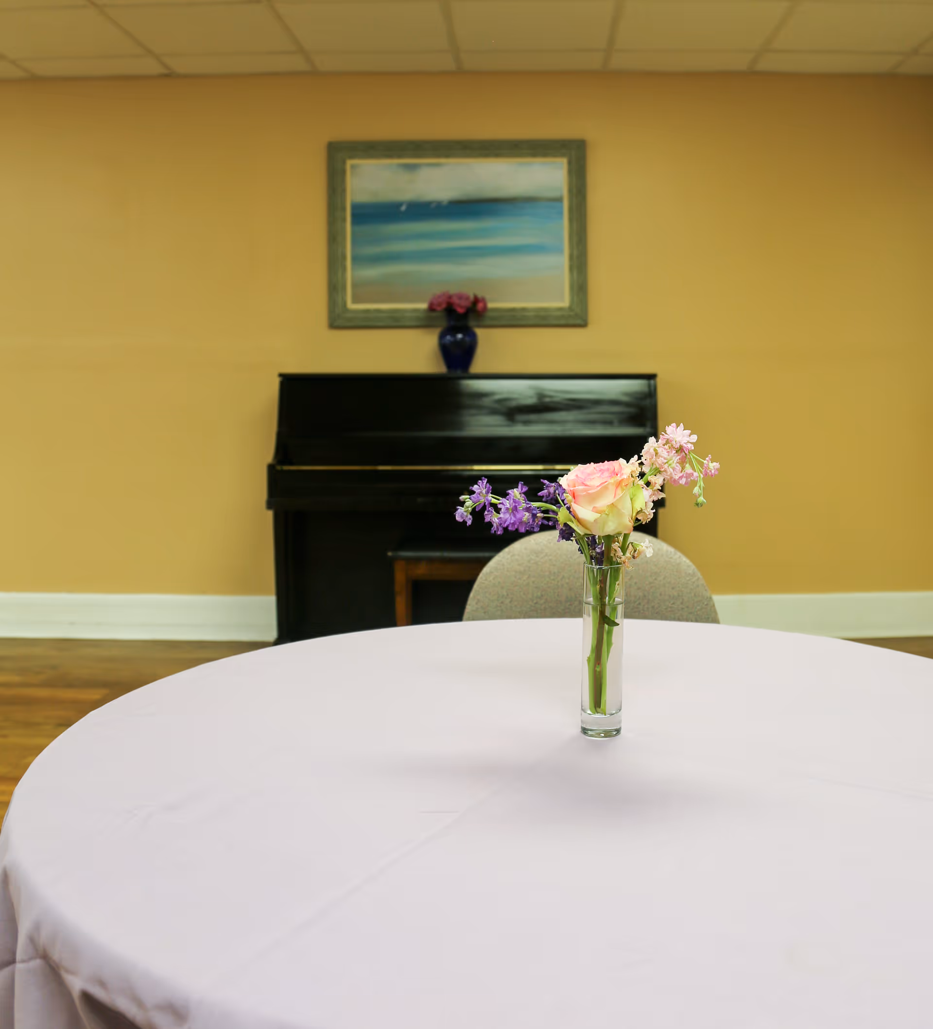A round table covered with a white tablecloth with a small glass vase holding a bouquet of flowers in the center. Behind the table is a chair and a black upright piano with a blue vase holding flowers on top. Above the piano hangs a framed painting of a seascape.