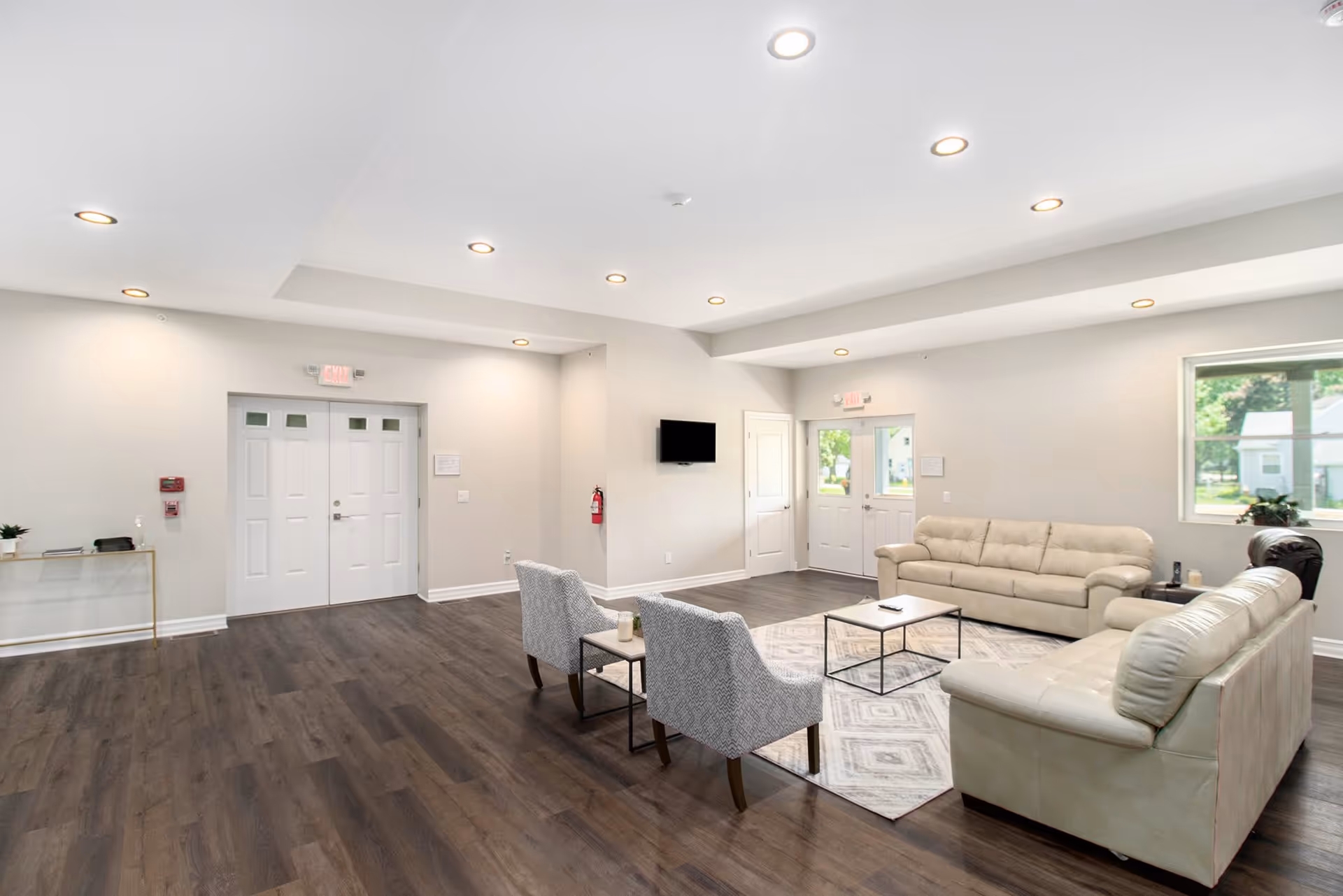 A spacious, well-lit living room area with light-colored walls and dark wood flooring. The room features two beige leather sofas, two patterned armchairs, a rectangular coffee table on a patterned rug, a wall-mounted TV, and large windows letting in natural light. There are two sets of double doors and a fire extinguisher mounted on the wall.