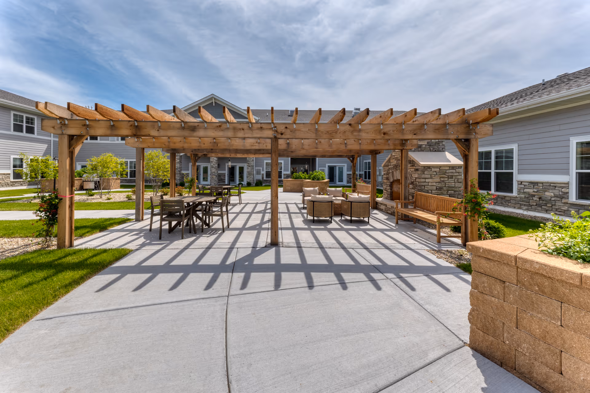 Outdoor patio area at Cedarhurst Senior Living of McHenry featuring a wooden pergola casting shadows on a concrete surface. The space includes several seating arrangements with chairs and tables, a stone fireplace, and surrounding greenery with bushes and grass. The building exterior is visible in the background under a partly cloudy sky.