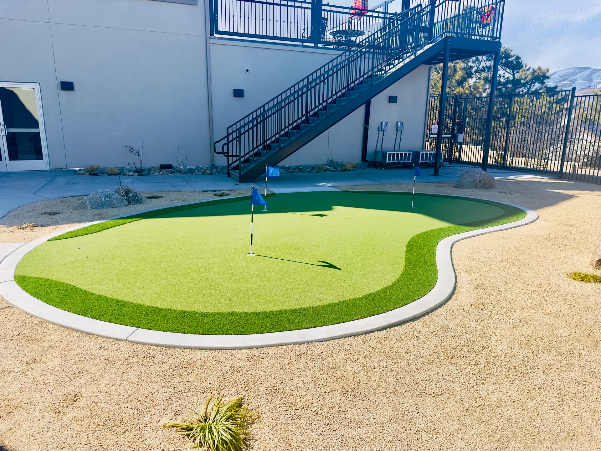 Outdoor putting green with three small flags on a synthetic turf surface, surrounded by gravel and landscaping. In the background, there is a building with a staircase leading to an upper level and a fenced area with trees and mountains visible in the distance.