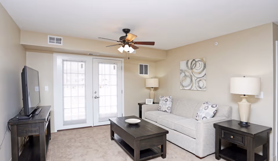 Bright furnished living room with a beige sofa, coffee table, TV on a stand, lamps, and French doors.