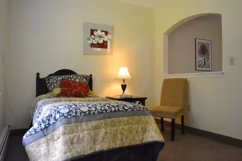 A cozy bedroom with a single bed covered in a patterned quilt and multiple pillows. A small bedside table with a lamp and a book is next to the bed. There is a mustard-colored chair against the wall and a framed picture hanging above it. The walls are painted light yellow.