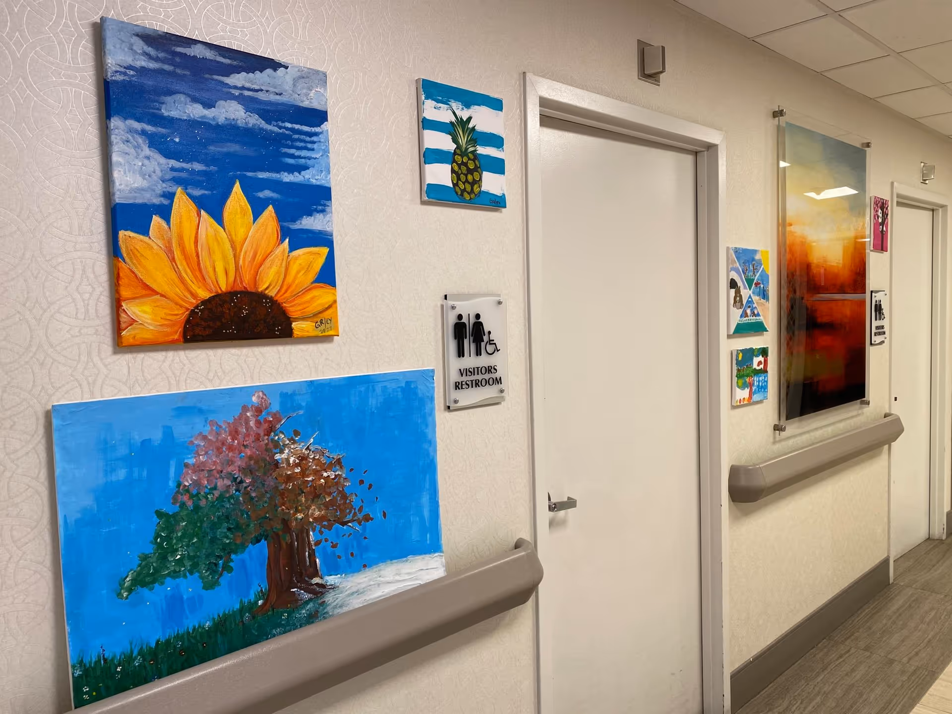 A hallway in a care center with several colorful paintings on the walls, including a sunflower, a pineapple, and a tree. There are two white doors with signs indicating a visitors restroom and a restroom for men and women. The walls have a light patterned wallpaper and there are handrails along the hallway.