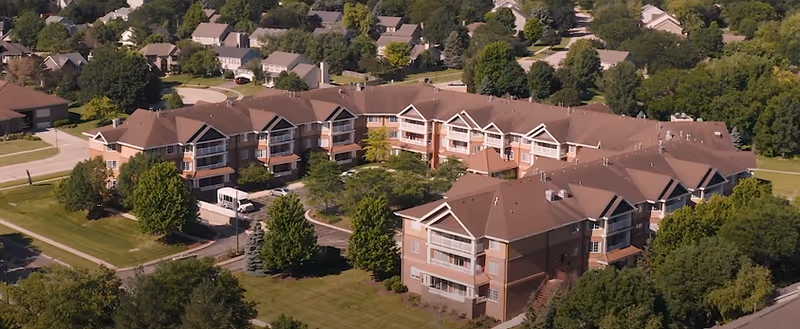 Aerial view of The Reserve of Geneva, a multi-story senior living facility surrounded by trees and residential houses, with well-maintained lawns and a driveway with a parked vehicle.