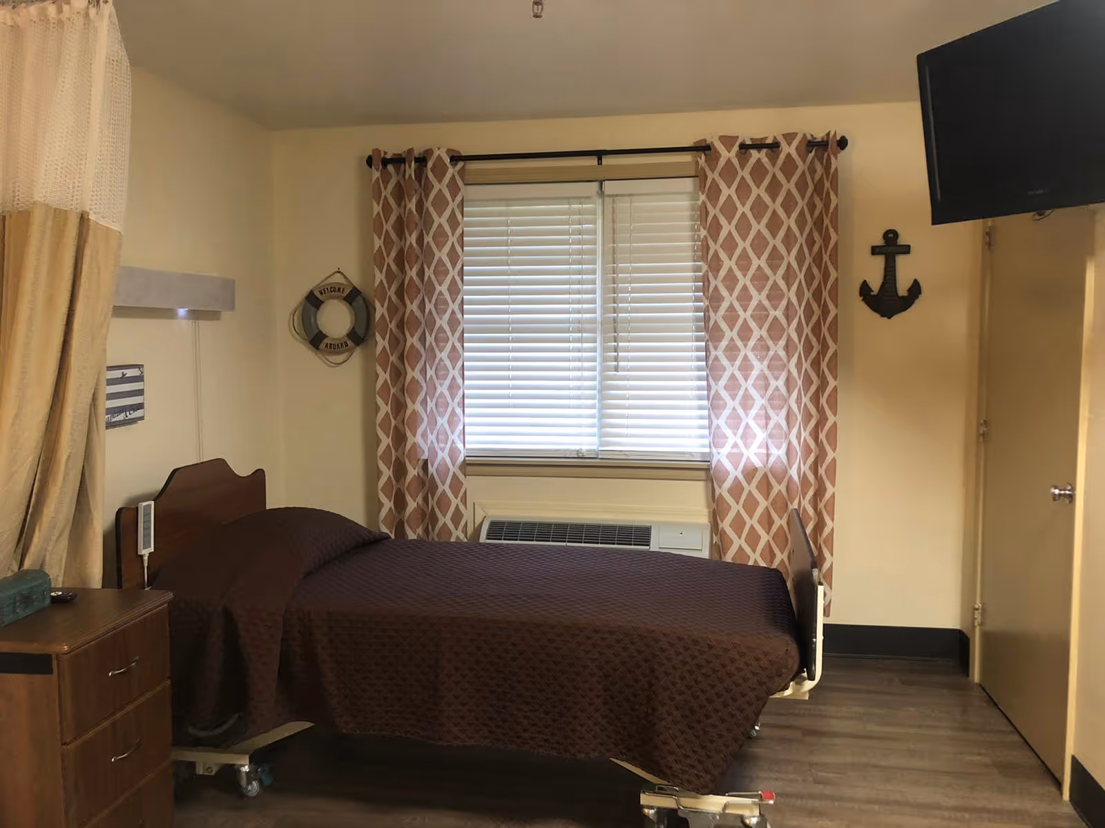 A small bedroom with a single hospital-style bed covered with a brown quilted blanket. There is a wooden nightstand with drawers next to the bed. The window has white blinds and patterned curtains in beige and white. On the wall, there is a decorative life preserver with the words 'Welcome Aboard' and a black anchor decoration. A flat-screen TV is mounted on the wall in the upper right corner.