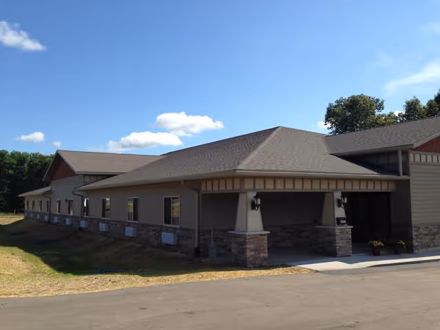 Exterior view of a single-story building with beige siding and stone accents under a clear blue sky with a few clouds. The building has multiple windows and a covered entrance with two wall-mounted lights.