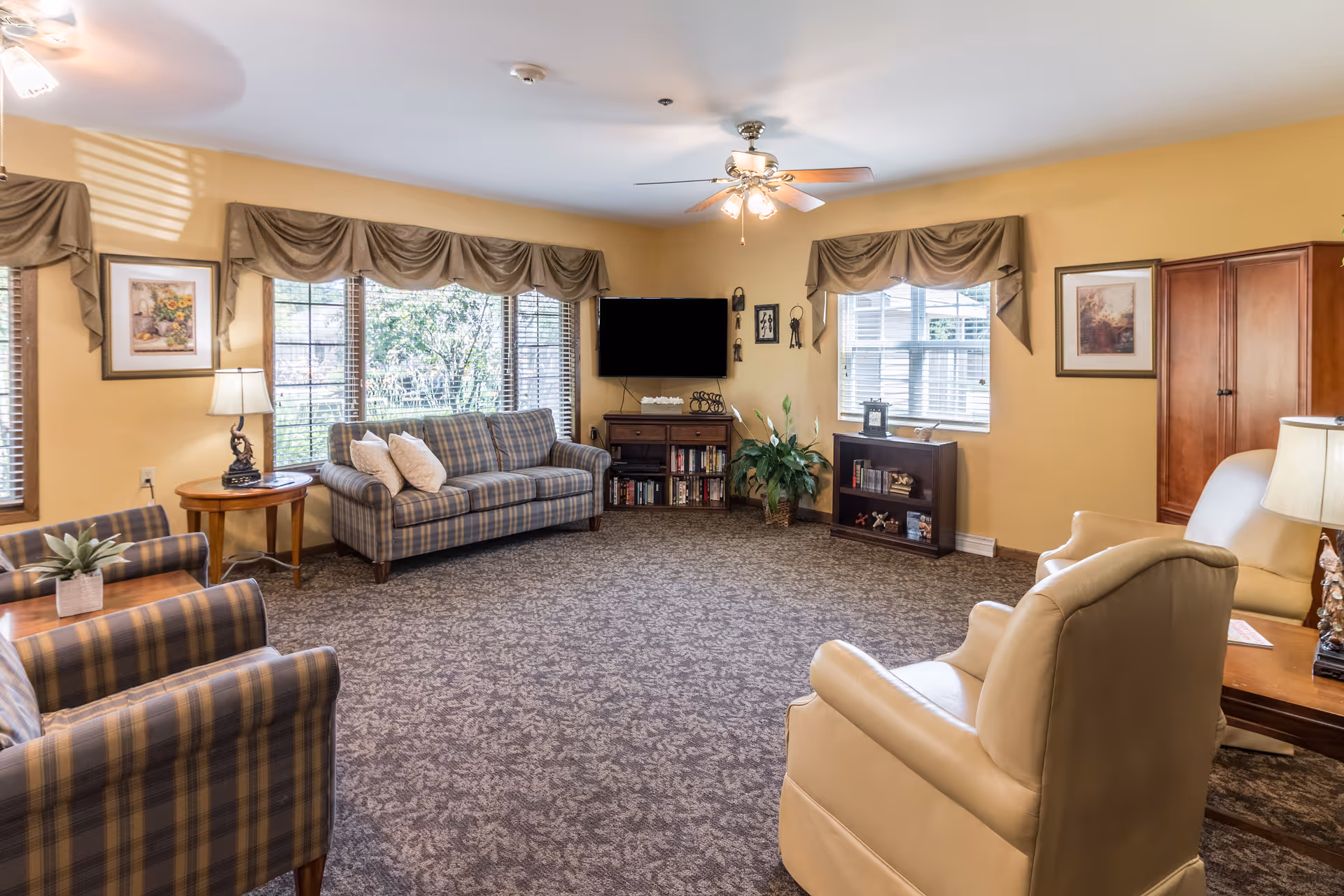 A cozy senior living room with beige walls and patterned carpet. The room features a plaid sofa and matching armchairs, a beige leather armchair, a wooden side table with a lamp, a ceiling fan with lights, a flat-screen TV mounted on the wall, bookshelves with books and decorative items, framed artwork on the walls, and windows with valance curtains letting in natural light.
