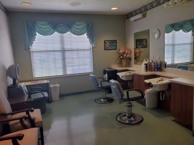 Interior of a salon room with two salon chairs in front of sinks and mirrors, a window with blue valance curtains, a small table with a hair dryer, and a few chairs along the wall.