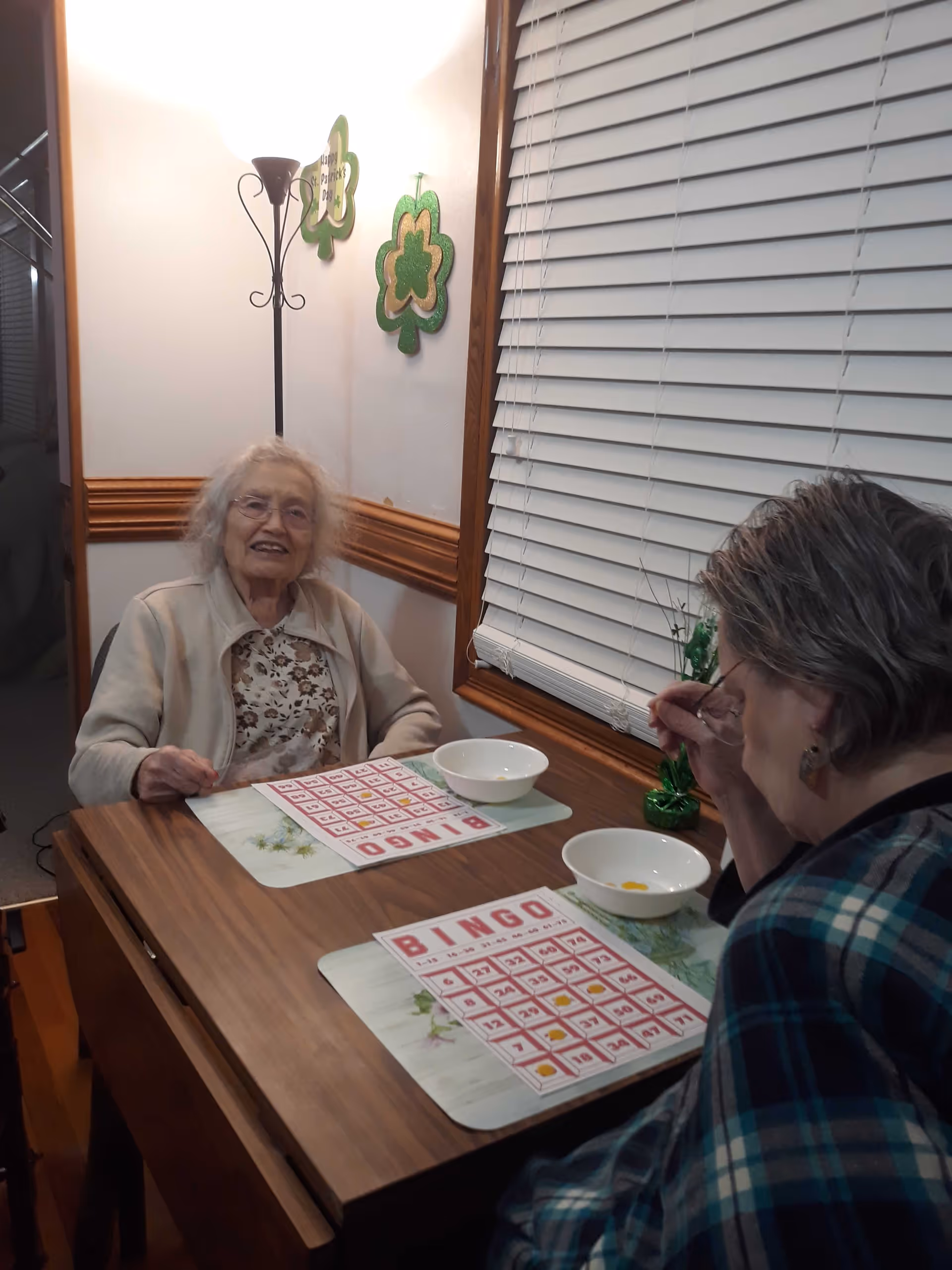 Two elderly women sitting at a wooden table playing bingo. One woman is smiling and looking at the camera, while the other is focused on her bingo card. There are two white bowls on the table and green shamrock decorations on the wall behind them.