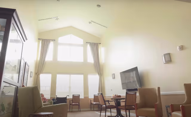 A bright living room area with high ceilings and large windows covered with curtains. The room contains several chairs arranged around small tables, a flat-screen TV mounted on the wall, and a glass display cabinet on the left side. The walls are painted light yellow, and the room has a calm, welcoming atmosphere.