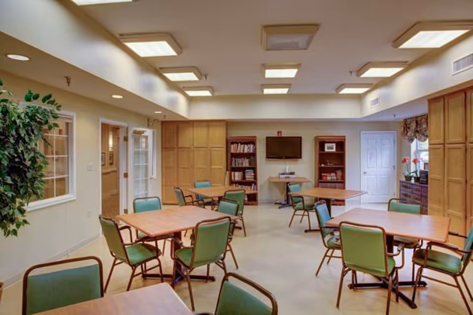 A well-lit common room with several square wooden tables surrounded by green cushioned chairs. The room features built-in wooden cabinets, bookshelves filled with books, a wall-mounted flat screen TV, and a small table with a device beneath the TV. There are windows and a door on the right side, with floral curtains and potted plants adding a touch of decor.
