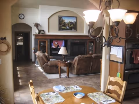 Open-plan dining area with a wooden table and chandelier opening into a living room with brown sofas and an entertainment center.