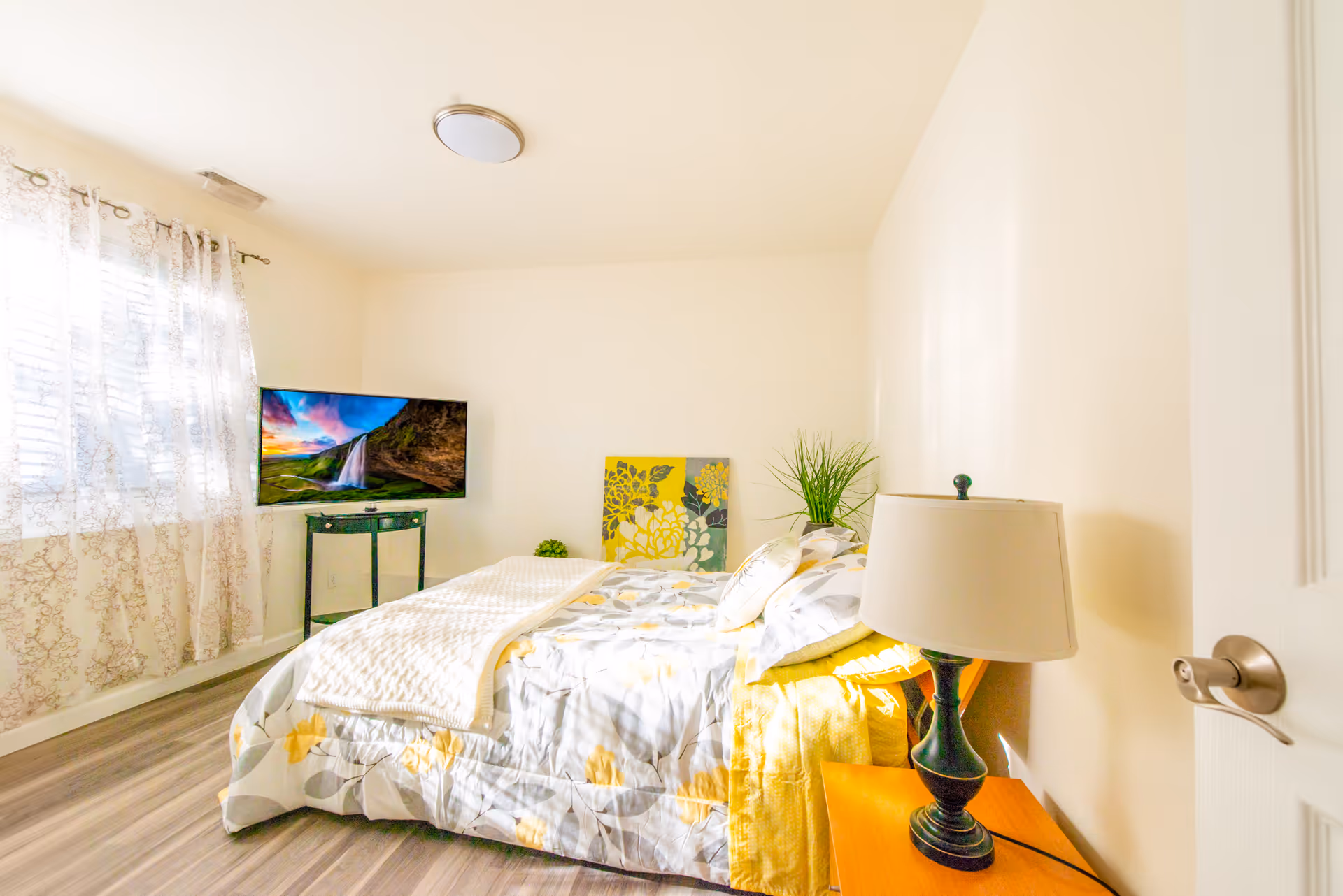 Sunlit bedroom with a made bed featuring yellow-gray bedding, a bedside lamp on a wooden table, a TV on a stand, and a window with patterned curtains.