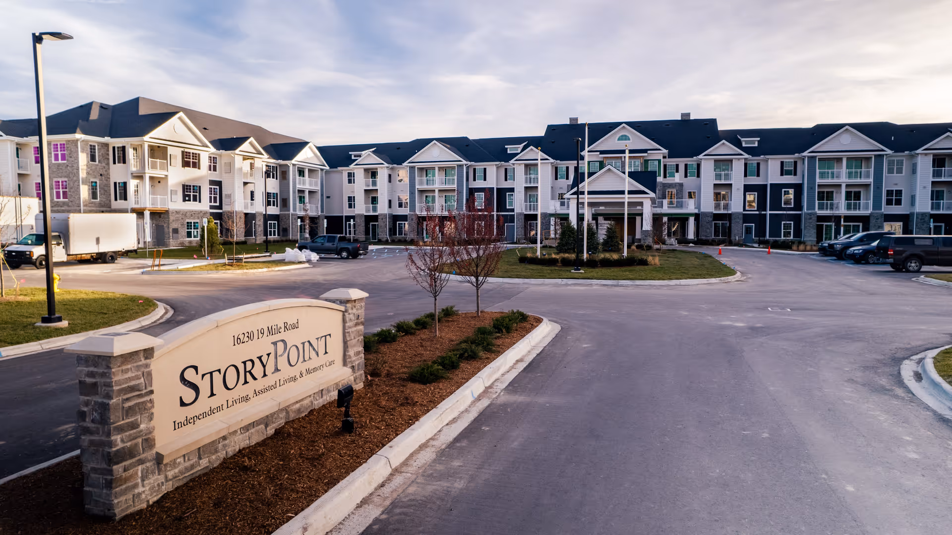 Exterior view of StoryPoint Clinton Township senior living facility showing a large three-story building with multiple balconies and windows, a driveway with parked vehicles, landscaped areas with small trees and shrubs, and a stone sign at the entrance displaying the facility's name and address.