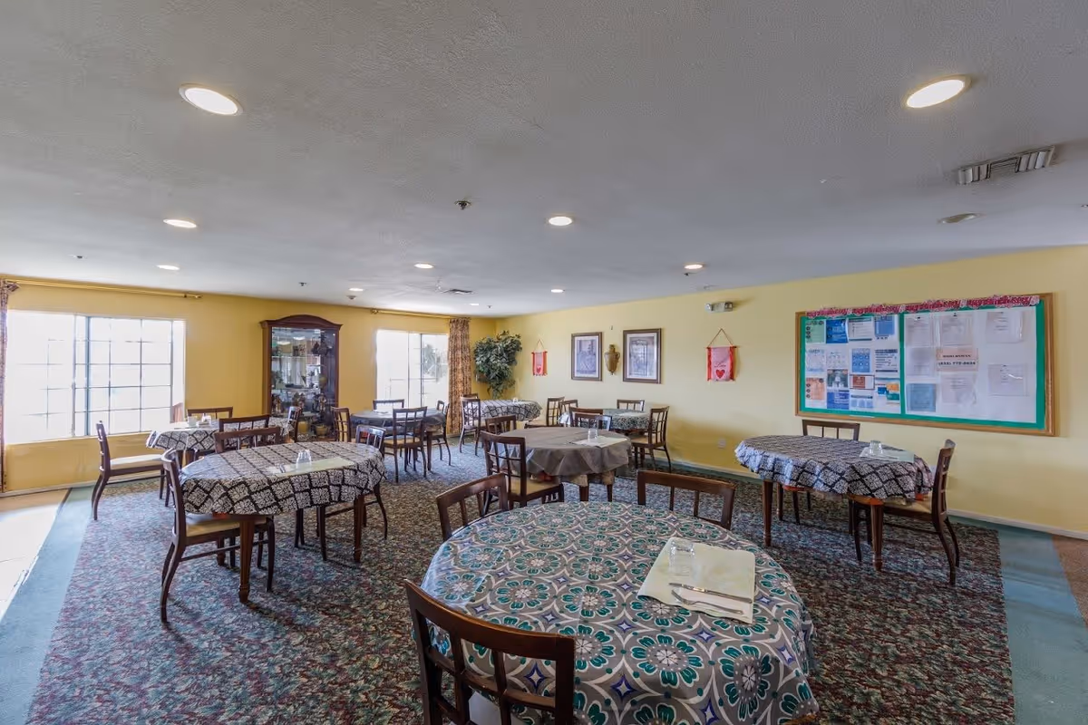 A dining room with multiple round tables covered with patterned tablecloths and wooden chairs around them. The room has yellow walls, two large windows letting in natural light, a bulletin board with various notices, framed pictures on the wall, and a glass display cabinet in the corner.