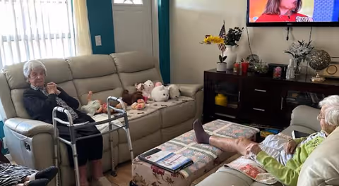 Two elderly women sitting in a living room on beige sofas. One woman is sitting near a walker, and the other has her legs resting on a floral ottoman. A TV is mounted on the wall above a dark wooden cabinet with decorative items and flowers.