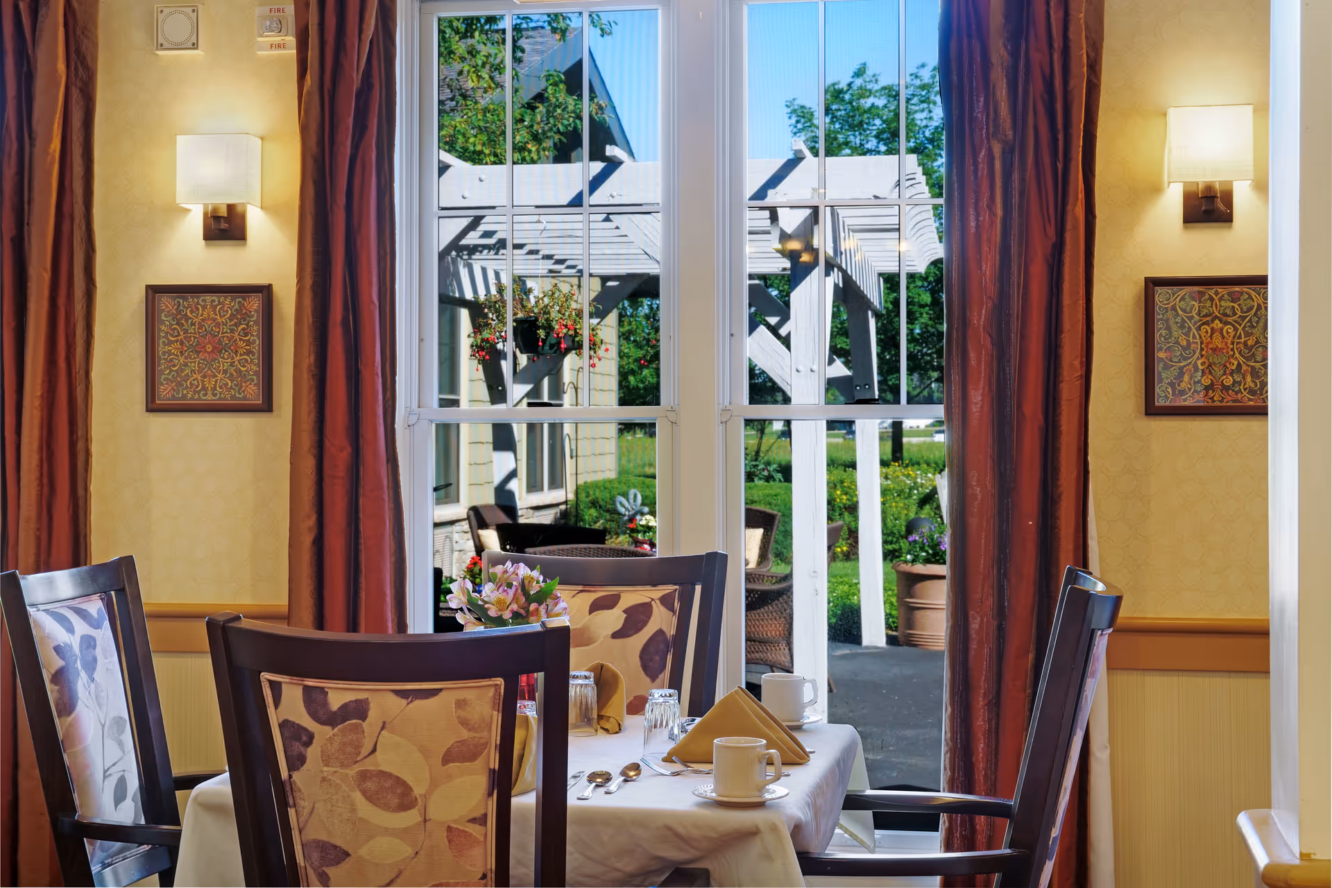 Sunlit dining table set with chairs and place settings beside large windows looking out to a pergola and garden.