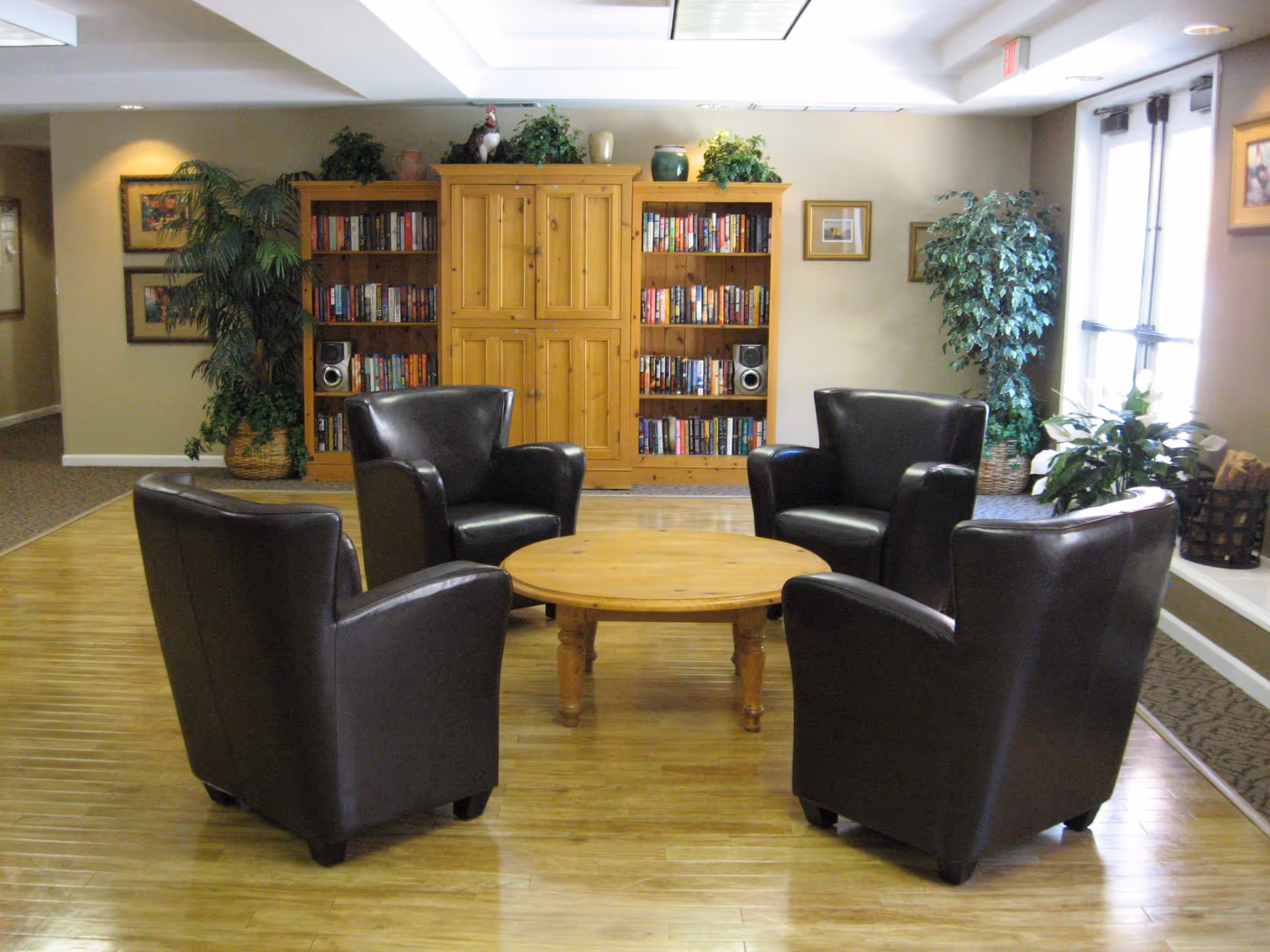 A cozy sitting area with four black leather armchairs arranged around a round wooden coffee table. Behind the chairs is a wooden bookshelf filled with books and decorative plants on top. The room has wooden flooring, framed pictures on the walls, and large windows letting in natural light.
