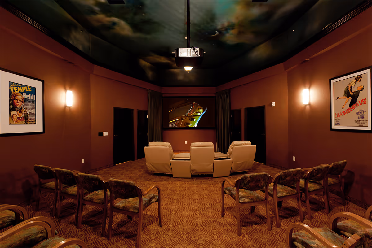 Community movie theater room with rows of upholstered chairs and three recliners facing a projection screen under a painted ceiling.