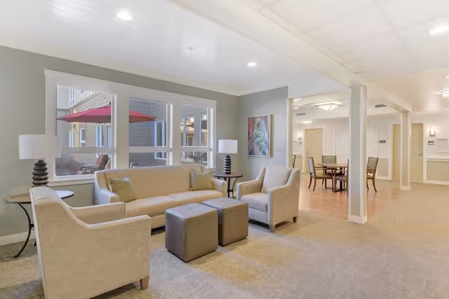 A bright and spacious living room area in a senior living facility featuring a beige sofa, two matching armchairs, two small square ottomans, two side tables with lamps, and large windows showing an outdoor patio with a red umbrella. In the background, there is a dining area with a table and chairs, light-colored walls, and ceiling lights.