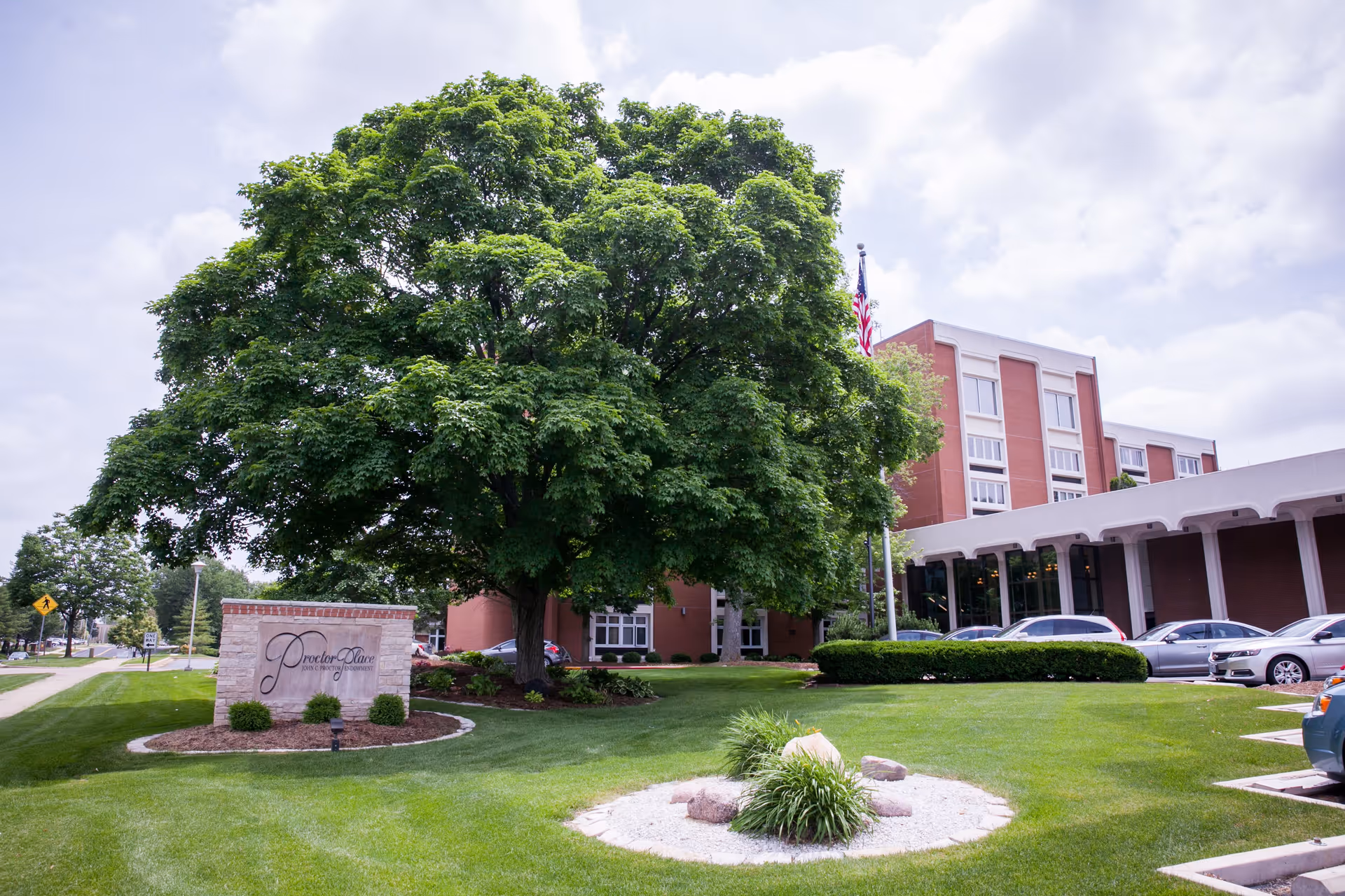 Exterior view of Proctor Place Senior Living facility showing a large green tree, well-maintained lawn, a stone sign with the facility name, parked cars, and a multi-story brick building under a partly cloudy sky.