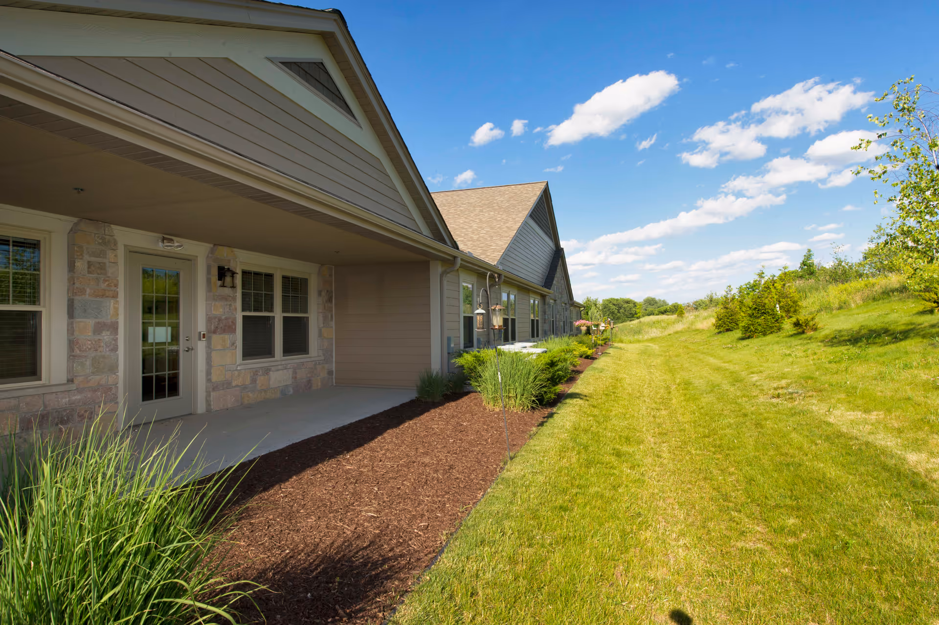Exterior view of a single-story building with stone and siding walls, a covered patio area, and a well-maintained lawn and garden beds under a blue sky with scattered clouds.