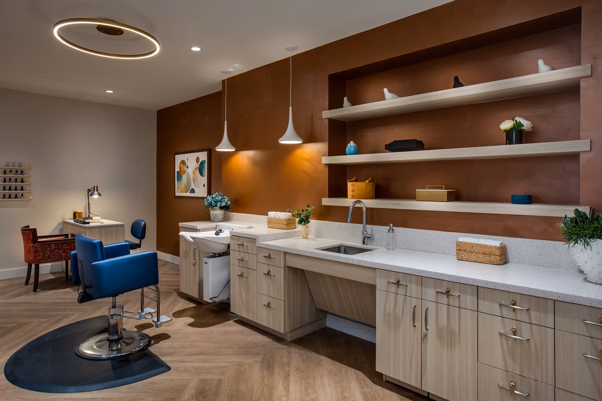 Well-lit salon interior with a countertop and sink, open shelves and cabinetry, two blue styling chairs, and a manicure desk.