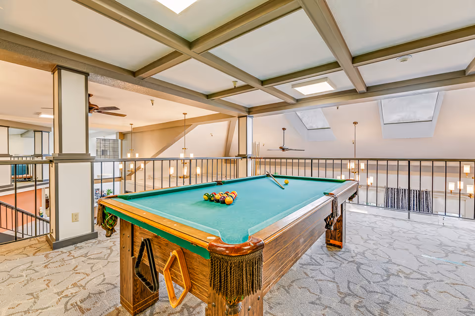 A loft-style indoor recreation area with a billiards table on a carpeted floor overlooking a lower level through metal railings and skylights.