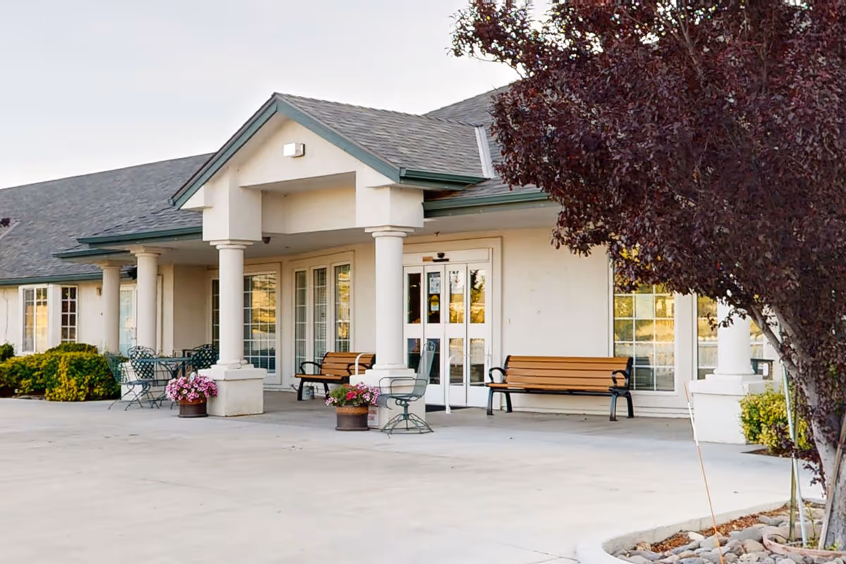 Exterior view of a senior living facility entrance with white columns, benches, potted flowers, and a tree with dark red leaves on the right side.