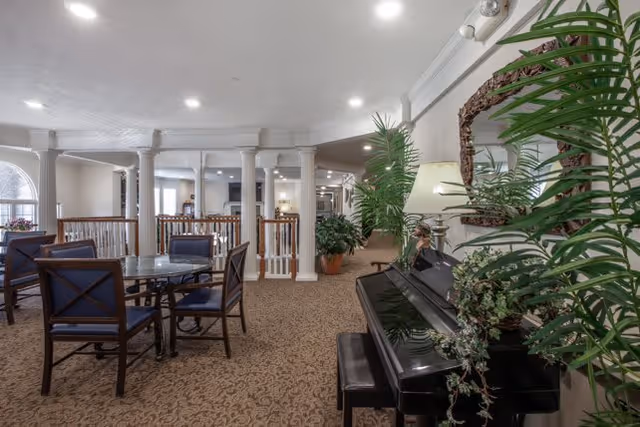 Interior view of a senior living facility lounge area with a black piano adorned with plants and a lamp on the right side, several round glass tables with chairs, carpeted floor, white columns, and soft overhead lighting.