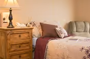 A cozy bedroom featuring a made bed with decorative pillows, a wooden nightstand with a lamp, and an upholstered chair.