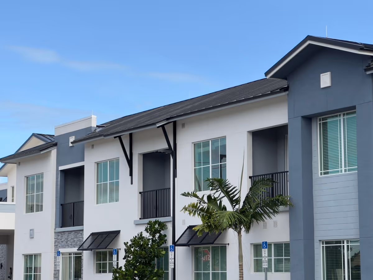Front exterior of a two-story modern senior living building with balconies, large windows, and palm trees.