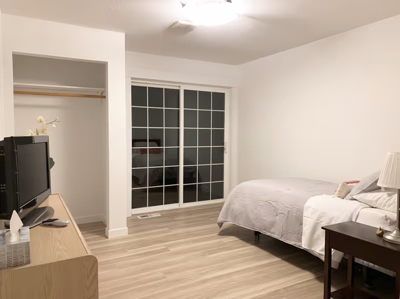Minimally furnished bedroom with a bed and nightstand on the right, a TV on a dresser to the left, an open closet, and sliding glass doors straight ahead.
