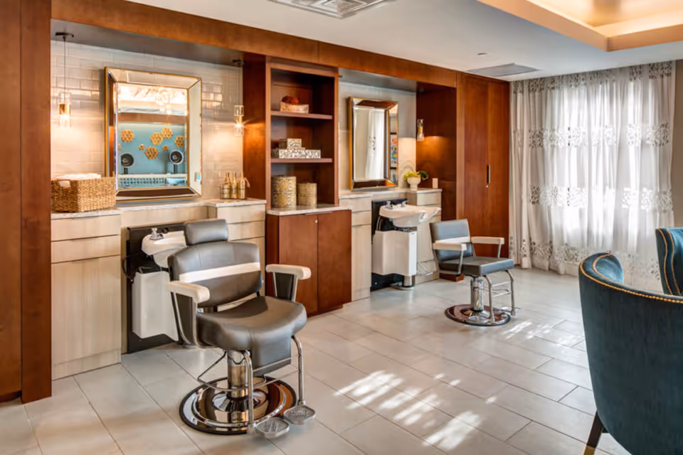 Interior view of a salon area with two gray salon chairs in front of washbasins and mirrors, wooden cabinetry, and shelves with decorative items. The room has large windows with sheer white curtains allowing natural light to fill the space, and a blue upholstered chair is partially visible on the right.