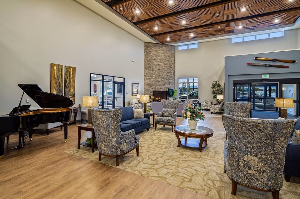 A spacious senior living facility common area with a high ceiling and wooden floor. The room features a grand piano on the left, several upholstered armchairs and sofas arranged around a round glass coffee table with a flower vase. There is a stone fireplace in the background, large windows, and decorative wall art including two paintings and a wooden propeller above the entrance doors.