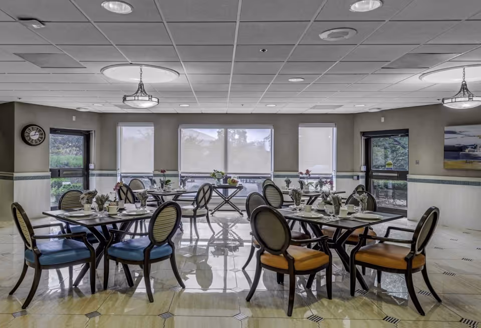 A dining room with several tables set for meals, each table has chairs with different colored cushions, place settings, and small flower arrangements. The room has large windows with blinds, two doors leading outside, and a clock on one wall.