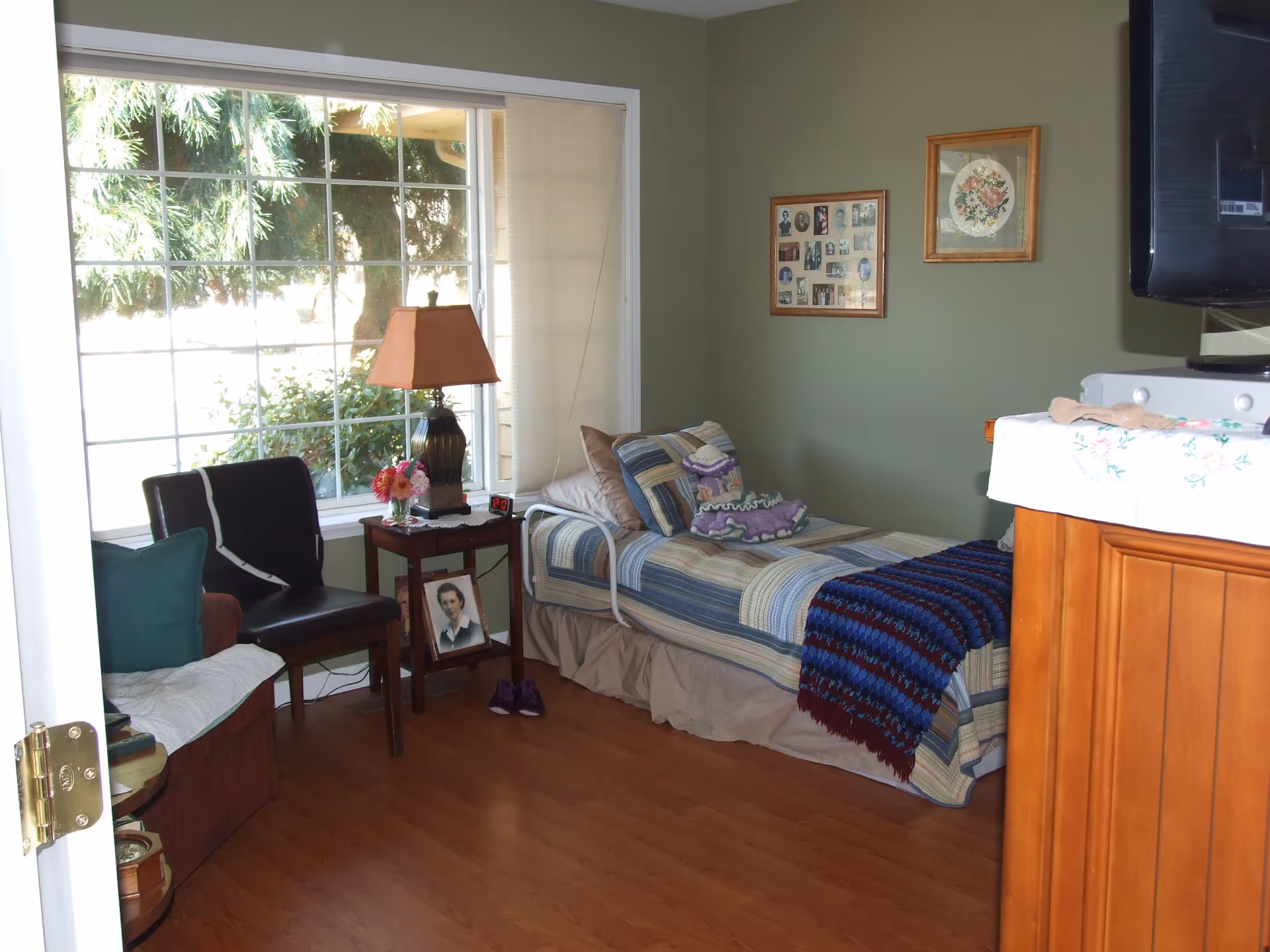 A cozy bedroom with a single bed covered in striped bedding and a blue knitted blanket. There is a small wooden side table next to the bed with a lamp, flowers, and a framed photo underneath. Two chairs, one with a green pillow, are placed near a large window with a view of greenery outside. The walls are painted green and decorated with framed pictures. A TV is mounted on the wall above a wooden cabinet with a floral cloth on top.