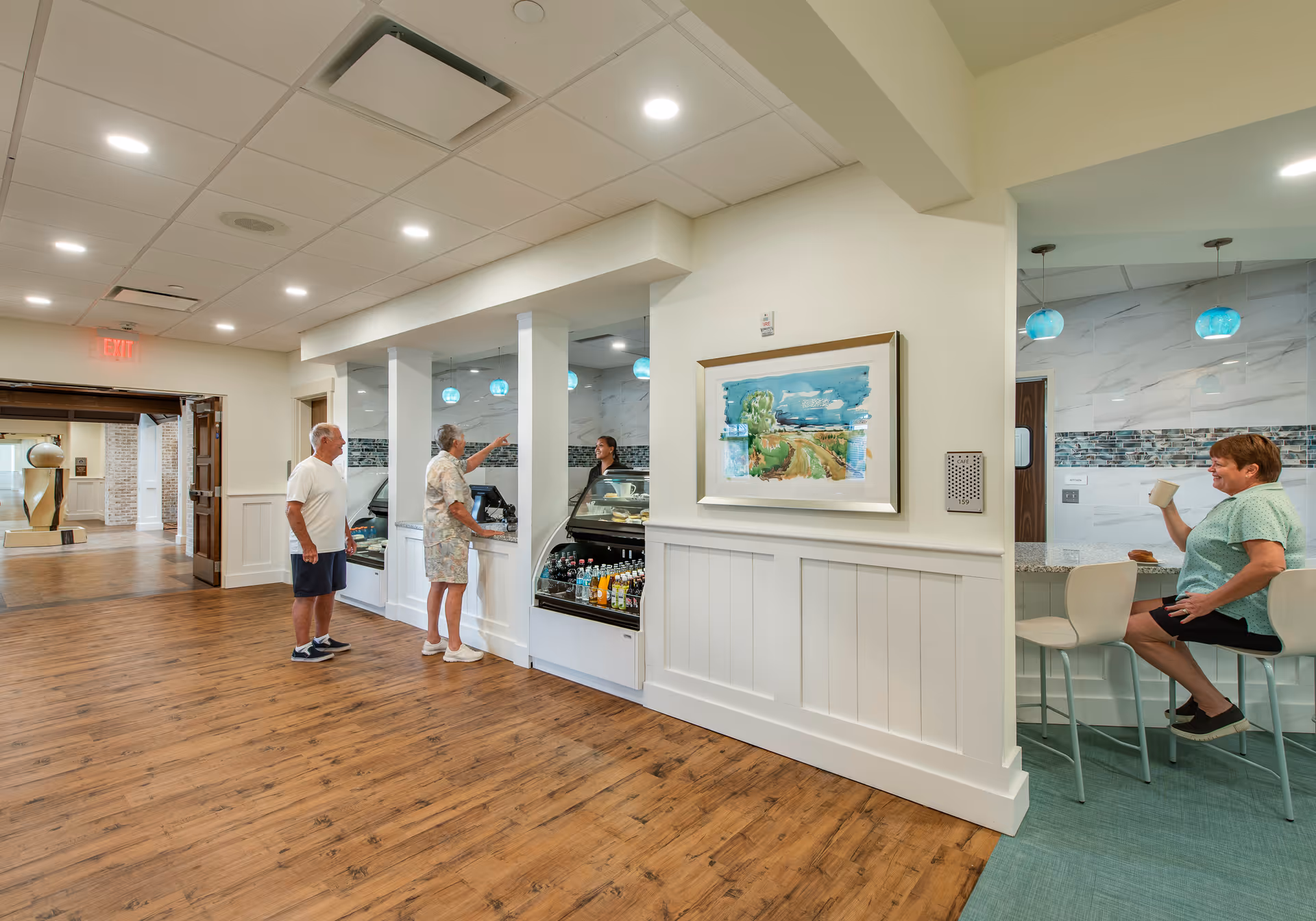 Interior view of a senior living facility showing a small cafe or snack bar area with two elderly people standing at the counter interacting with a staff member behind the glass display. Another elderly woman is sitting on a bar stool to the right, drinking from a mug. The area has wood flooring, white walls with wainscoting, and modern lighting fixtures.