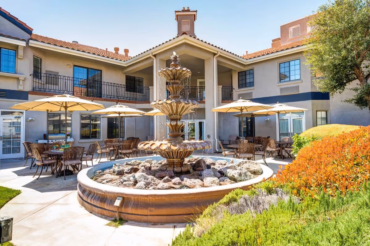 Outdoor courtyard area of Whispering Winds of Apple Valley featuring a multi-tiered stone fountain in the center surrounded by patio tables with umbrellas and chairs. The building has two stories with balconies and large windows, and there are landscaped bushes and greenery around the courtyard.