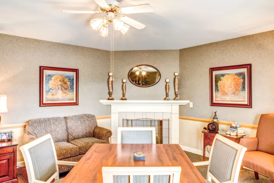 A cozy living room with a wooden table surrounded by four chairs in the foreground. Behind the table is a patterned loveseat, a white fireplace with decorative candle holders and an oval mirror above it. On either side of the fireplace are framed pictures of trees with autumn foliage. To the right is an orange armchair next to a small wooden side table with books and a decorative clock. The ceiling has a white fan with lights.