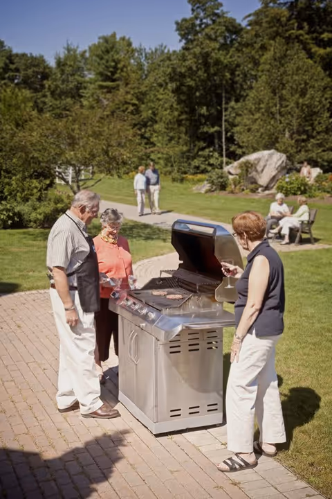 Three elderly people standing around a stainless steel outdoor grill on a paved pathway in a garden setting. One man is grilling while two women stand nearby, one holding a glass of wine. In the background, two elderly people are walking on the path and two others are seated on a bench in a grassy area with trees and large rocks.