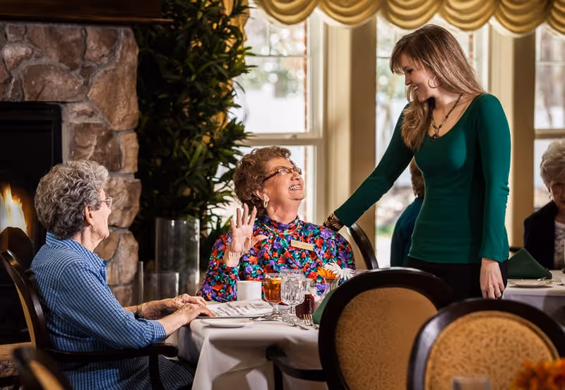 Two elderly women sitting at a dining table in a cozy room with a stone fireplace, engaged in conversation with a younger woman standing beside them, smiling. The table is set with glasses, cups, and a flower centerpiece, and large windows with yellow curtains are in the background.