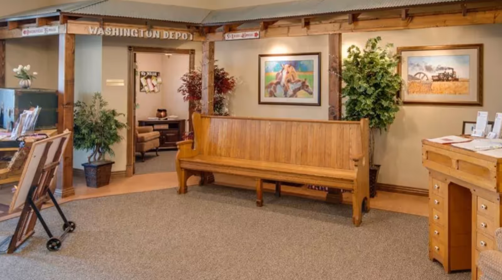 Interior view of a senior living facility lobby area with wooden bench seating, framed paintings on the walls, potted plants, and a wooden desk with brochures. A wooden beam structure above the entrance reads 'WASHINGTON DEPOT'.