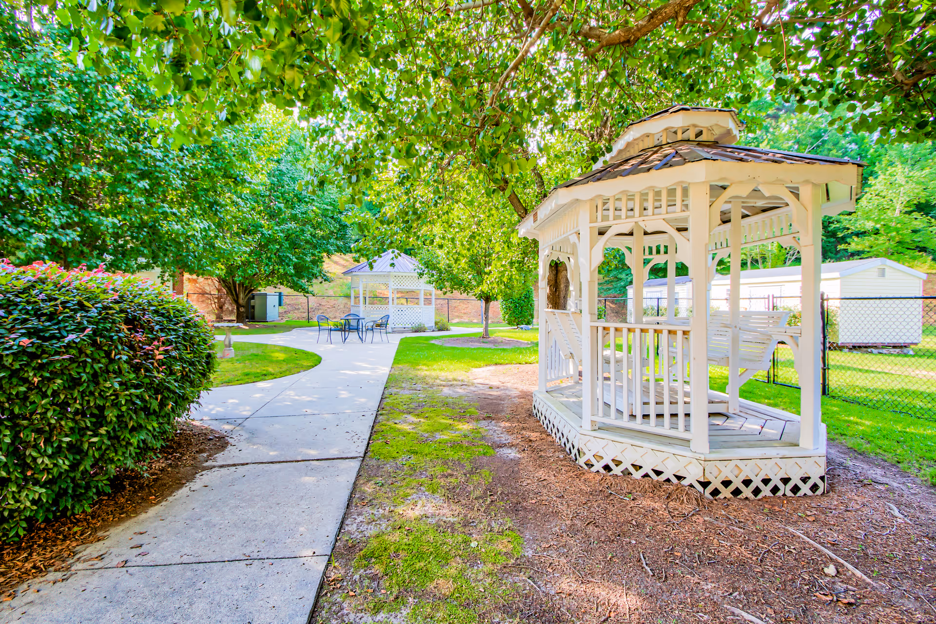 A bright outdoor garden area with a white wooden gazebo featuring a swing, surrounded by green trees and bushes. A paved walkway leads to another white gazebo with a table and chairs in the background, under a sunny sky.
