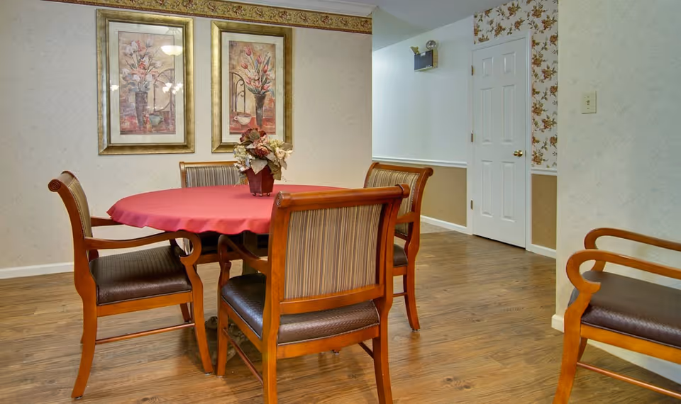 A dining area with a round wooden table covered with a red tablecloth and surrounded by five wooden chairs with cushioned seats and backs. A floral centerpiece is on the table. The room has wood flooring, floral wallpaper on one wall, and two framed floral paintings hanging on the wall.