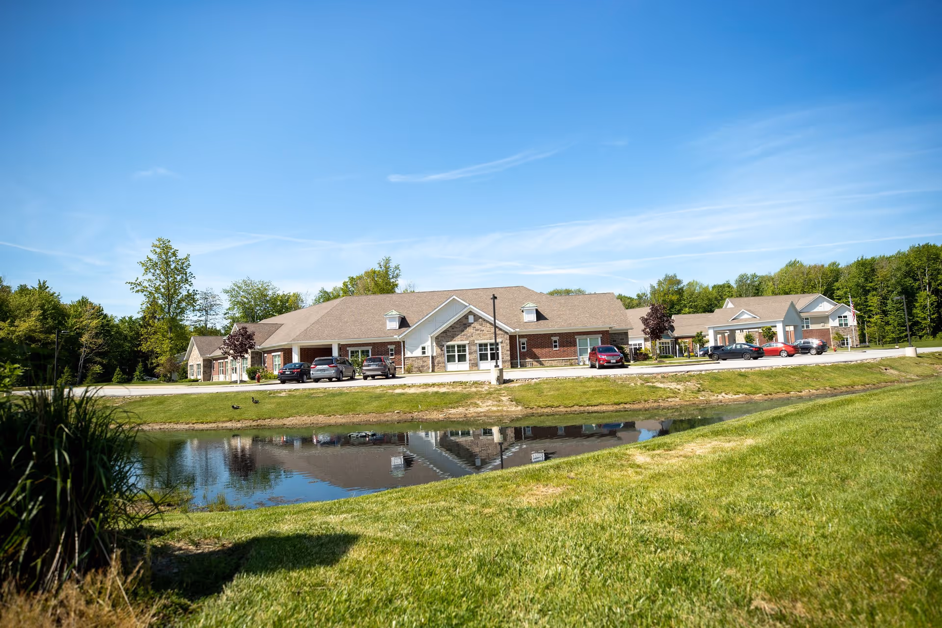 Exterior view of a single-story senior living facility building with a brick and stone facade, several parked cars in front, a small pond reflecting the building, and green grass and trees surrounding the area under a clear blue sky.