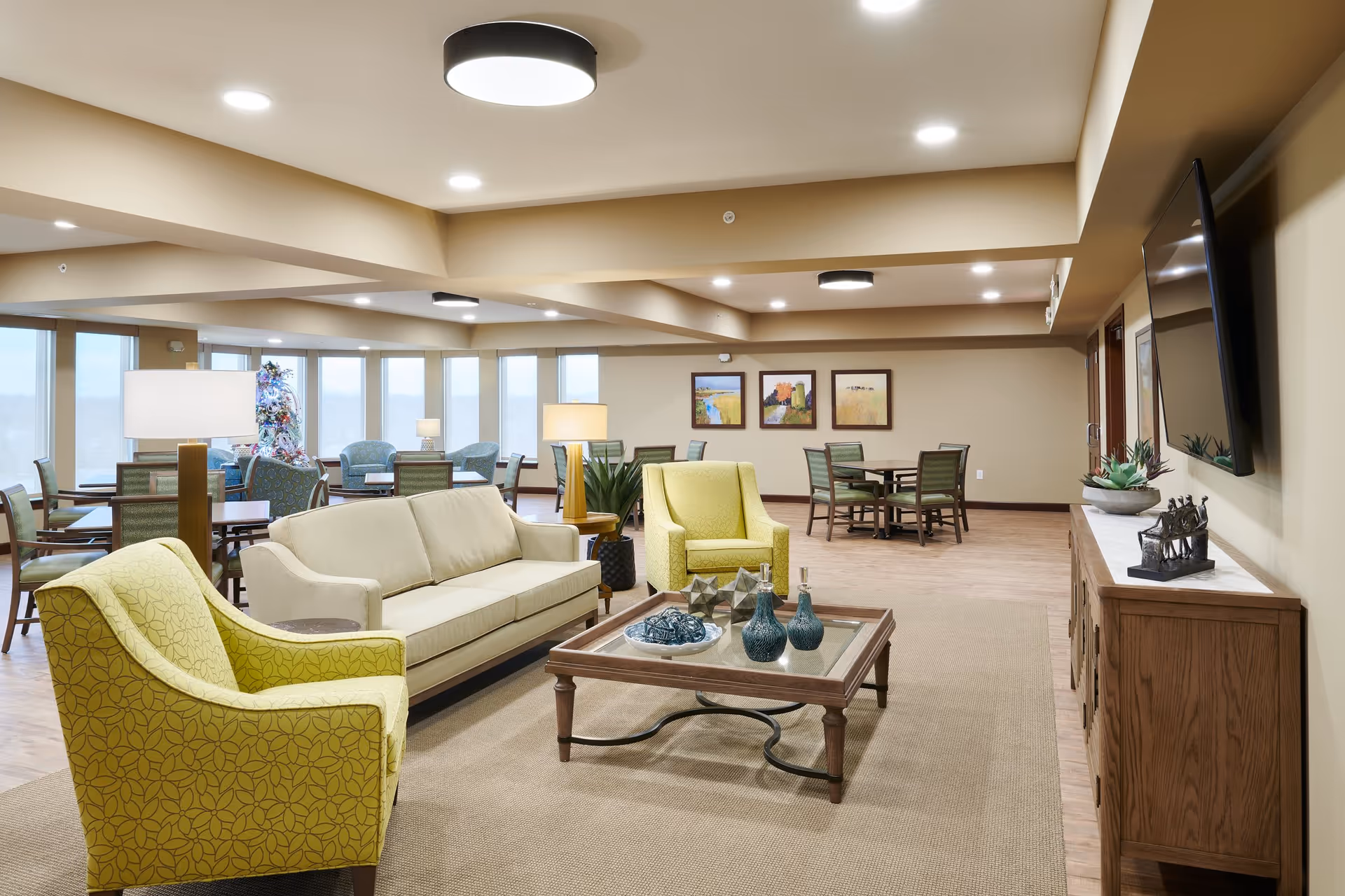 Spacious furnished senior living common area with sofas, armchairs, a coffee table, dining tables, and a wall-mounted TV near large windows.